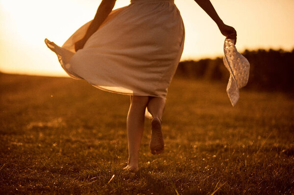 Girl in white dress barefoot running across the field on sunset background.