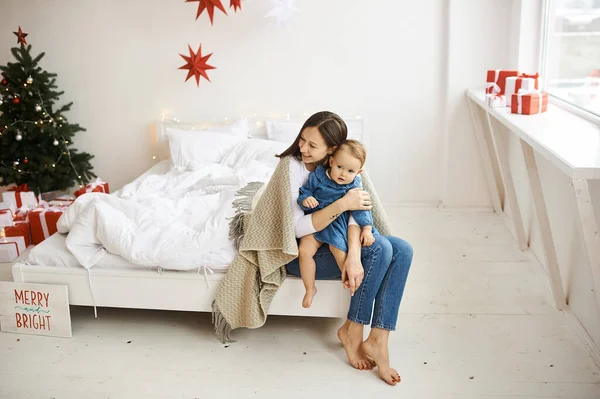 Счастливая мать, покрытая одеялом, и ее дочь, сидящая на краю кровати в светлой комнате. — стоковое фото