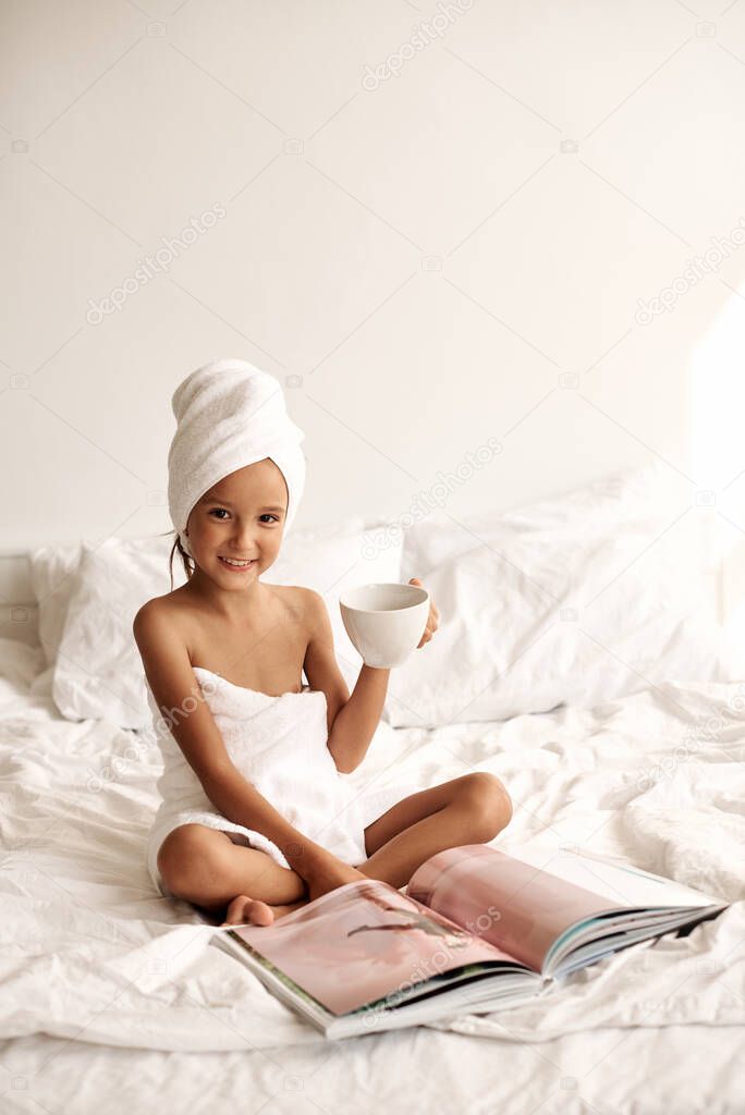 Fashion portrait of a little girl wrapped in a bath towel with a cup of milk in her hand reading a magazine. Child has fun in the morning sitting on the bed of a bright hotel room.