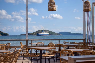 Güneşli bir Yunan adasının kıyısındaki resim gibi bir sahil restoranı. Bir feribot arka plandaki küçük yeşil adaları geçerek okyanusta yelken açıyor. Avrupa plajında akşam yemeği yerleri.