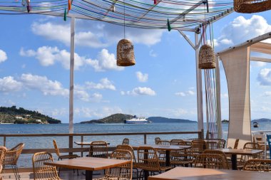 Güneşli bir Yunan adasının kıyısındaki resim gibi bir sahil restoranı. Bir feribot arka plandaki küçük yeşil adaları geçerek okyanusta yelken açıyor. Avrupa plajında akşam yemeği yerleri.