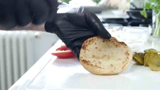 Человек на кухне раздает майонез с нарезанными огурцами на половине булочки за гамбургер — стоковое видео