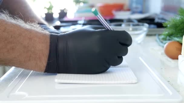 Close-up schieten met een persoon in de keuken dragen van zwarte handschoenen in handen schrijven van een koken recept — Stockvideo