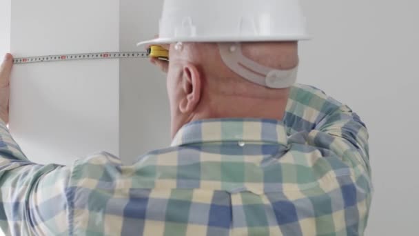 Un ingénieur portant un casque blanc utilise un ruban à mesurer et prend la dimension du mur dans une pièce d'appartement — Video