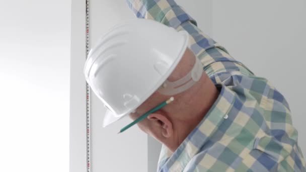 Un travailleur portant un casque blanc utilise un ruban à mesurer et prend la dimension du mur dans une pièce d'appartement — Video