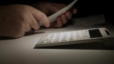 Man Working at Night, with Financial Documents, Makes Calculations Using Adding Machine. Fee and Taxes Calculated with Calculator.