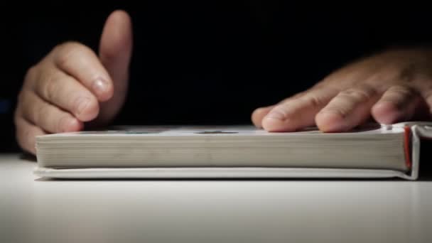 Homem vestindo óculos de leitura e aprendizagem tarde da noite a partir de um livro. uma mão que vira através das páginas de um livro aberto. — Vídeo de Stock