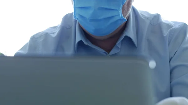Maskeli ve koruyucu eldivenli bir hastane ofisinde dizüstü bilgisayar kullanan bir doktor.