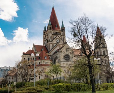 Wide view of a Romanesque church with red spires and stone architecture surrounded by trees and greenery under partly cloudy sky.