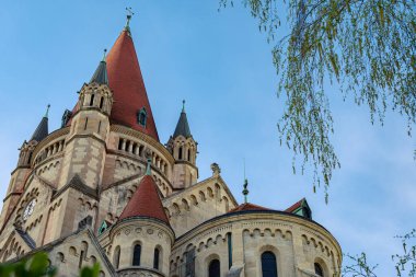 Close-up architectural detail of a historic Romanesque church with red tiled spires and ornate stone design under clear blue sky, captured from low angle.