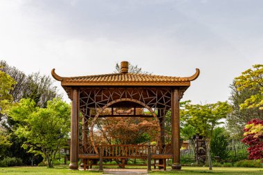 Traditional wooden Chinese-style gazebo surrounded by lush green trees in a peaceful garden setting.