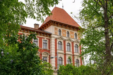 Historic European building with ornate architecture surrounded by lush green trees. The red brick facade, arched windows, and decorative roof details create a scene of classic design and heritage.