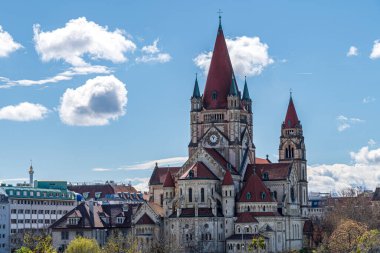 Viyana, Avusturya 'da bulutlu mavi gökyüzü altında kırmızı çatıları ve yüksek kuleleri olan tarihi kilise, Neo-Romanesk mimari ve şehir manzarasını gözler önüne seriyor..