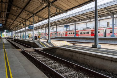 Parlak gün ışığında kırmızı banliyö trenleri ve metal çatıları olan boş tren istasyonu platformu, temiz mimari, demiryolu rayları ve ulaşım altyapısını gösteriyor.