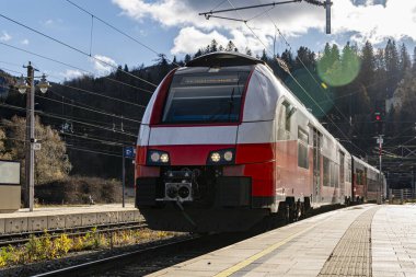 Gündüz vakti tren istasyonundaki modern kırmızı ve beyaz yolcu treninin arka planında dağlar ve ağaçlar seyahat, ulaşım ve bağlantıyı sembolize ediyor..