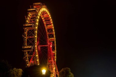 Parlak kırmızı ve sarı ışıklarda parlayan dönme dolap karanlık bir gece gökyüzüne karşı, sıcak tonlar ve kentsel gece atmosferiyle eğlenceli bir yolculuğun güzelliğini yakalıyor..
