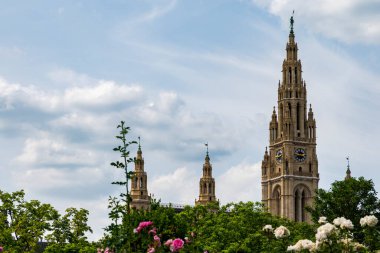 Yumuşak, bulutlu bir gökyüzünün altında yemyeşil bahçelerin üzerinde yükselen tarihi neo-gotik kuleler sakin ve zarif bir şehir manzarası yaratıyor..