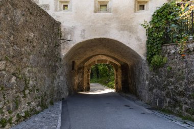 Eski taş geçit gölgeli bir yola çıkıyor, yıpranmış duvarlar, doğal dokular ve tarihsel bir kemerin içinden süzülen yumuşak güneş ışığı..