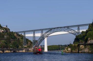 Geleneksel bir teknenin açık, güneşli bir günde aşağıda yelken açtığı manzaralı nehir manzarası boyunca uzanan çelik kemer köprüsü, seyahat, mimari ve Avrupa kıyısı güzelliğini gözler önüne seriyor..