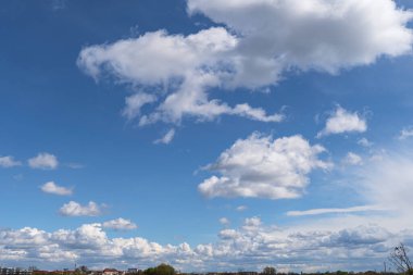 Uzak bir arazi üzerinde yumuşak beyaz kümülüs bulutlarıyla dolu parlak mavi gökyüzü doğa, hava ve arka plan temaları için ideal sakin ve açık hava atmosferini yakalıyor..