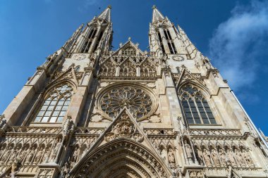 Büyük Gotik bir katedralin çarpıcı yükseliş manzarası. Detaylı taş oymalar, ikiz kuleler ve parlak mavi gökyüzüne bakan bir gül penceresi. Tarihi mimari ve görkemli dini tasarım..