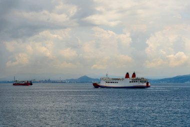 Yolcu feribotu bulutlu bir gökyüzü altında sakin kıyı sularında yol alıyor ve ufukta uzak tepeler var. Huzurlu deniz taşımacılığı sahnesi seyahat, lojistik ve günlük deniz geçişi..