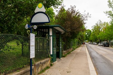 Ağaçlı bir cadde boyunca yeşil barınaklı sessiz bir banliyö otobüs durağı. Taze bahar yapraklı sakin şehir taşıma sahnesi, günlük gidiş-dönüş konsepti, huzurlu ve pratik ruh hali.