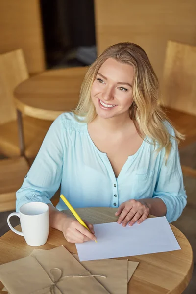 Beautiful young woman sitting at the table and writing letter looking to the side