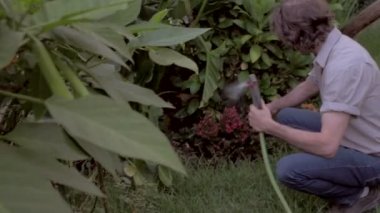 Orta yaşlı adam yavaş mo bir hortum yemyeşil bir açık tropikal Bahçe Sulama.