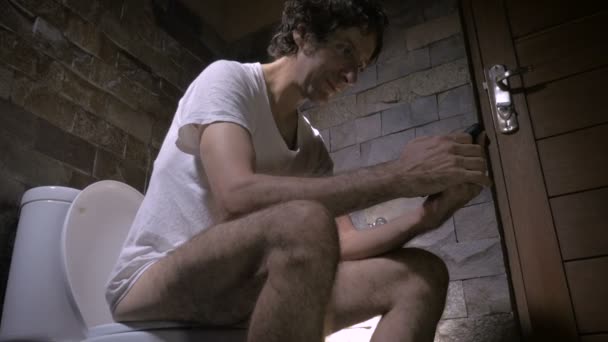 Low angle push-in of man sitting on a toilet laughing while on his cell phone — Stock Video