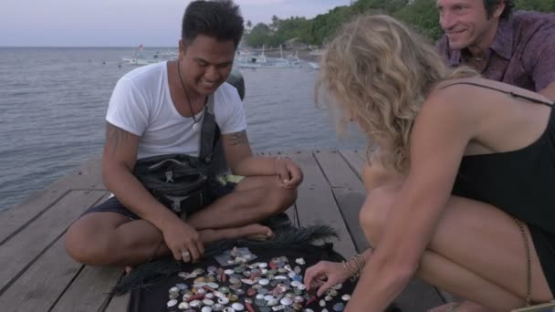 Two tourists buying jewelry from a vendor on a dock at dusk over the water — Stock Video