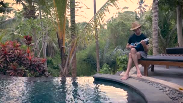 Amplio tiro de un hombre de mediana edad jugando un ukelele junto a la piscina en la selva — Vídeo de stock