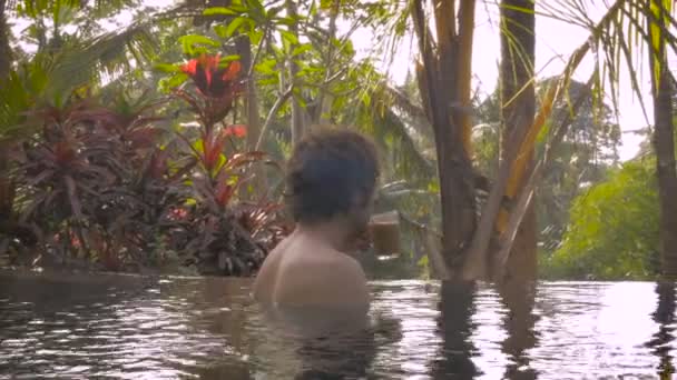 A man drinks coffee inside a pool in a gorgeous outdoor tropical island setting — Stock Video