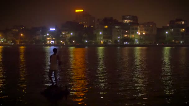 한 남자가 강물에 반사되는 도시의 불빛에 맞서 밤에 물고기를 잡습니다.. — 비디오