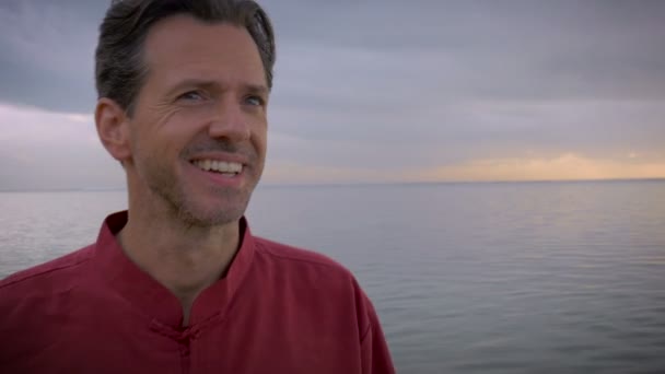 Handheld push-in: van middelste leeftijd man die lacht met uitzicht op het strand — Stockvideo