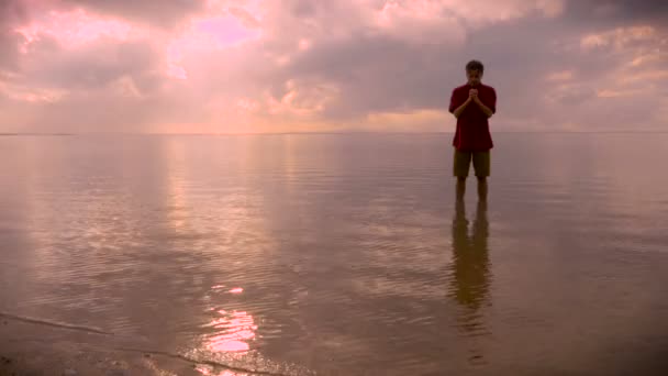 El hombre se encuentra en aguas tranquilas del océano rezando durante el amanecer — Vídeo de stock