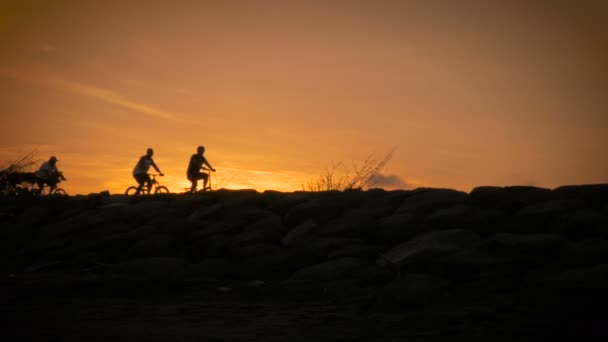 Siluetas de personas montando bicicletas y caminando en un paseo marítimo al atardecer — Vídeo de stock