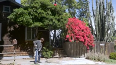 Bir adam evin önünde süpürgeyle garaj yolundan ölü yaprakları süpürüyor.