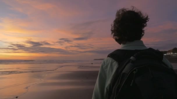 Un hombre camina con una mochila a lo largo de la playa durante el atardecer en slow mo — Vídeos de Stock