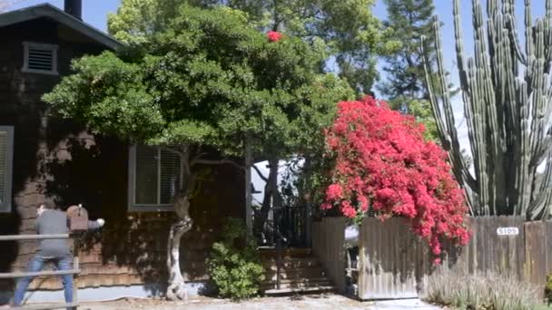 Человек в серой куртке подметает листья перед деревянным черепичным домом — стоковое видео