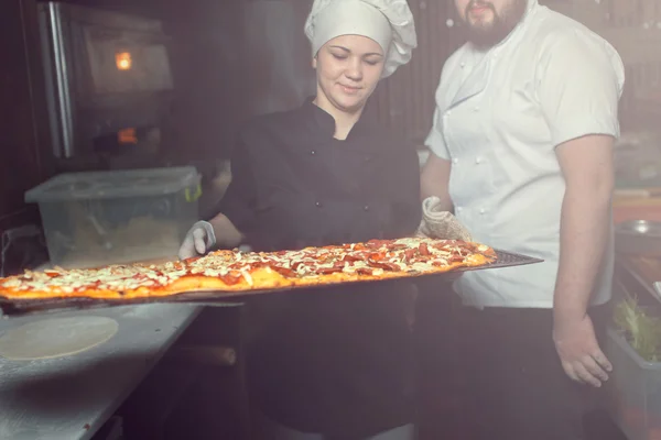 Повар-пекарь в черной форме кладет пиццу в духовку с лопатой на кухне ресторана — стоковое фото
