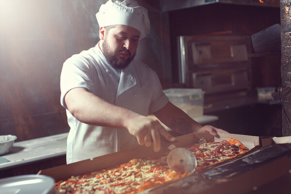 Closeup hand of chef baker in white uniform cutting pizza at kitchen