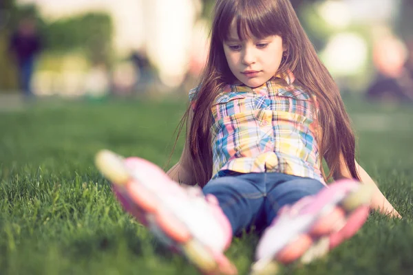 Flickan bär rullskridskor sitter på gräset i parken. — Stockfoto