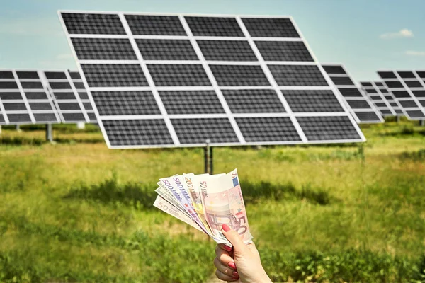 Hand holding money banknote with photovoltaic solar energy panels in background,