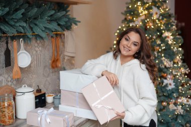 Gülümseyen kadın, arka planında süslü bir Noel ağacı olan bir mutfak ortamında hediye kutularını tutuyor..