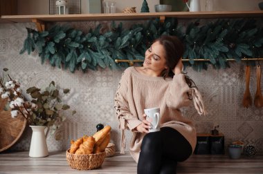 Elinde kupa tutan rahat kazaklı bir kadın, dekore edilmiş bir mutfakta kruvasan ve yeşillikle oturuyor..