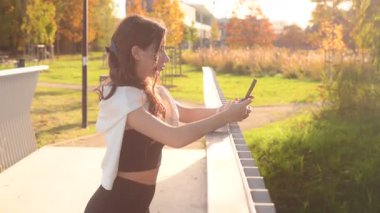 Açık hava eğitiminden sonra güneşli bir sonbahar gününde akıllı telefon kullanan aktif giyimli genç bir kadın.
