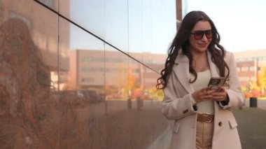 Güneş gözlüklü genç bir kadın, arka planda yansıtıcı bir bina görüntüsüyle şık bir bej ceket ve pantolon giymiş akıllı telefon kullanırken yürüyor.