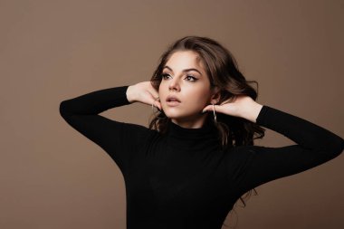 Siyah balıkçı yakalı genç bir kadının portresi kahverengi arka planda poz veriyor. Uzun kıvırcık saçlı bir mankenin fotoğrafı.