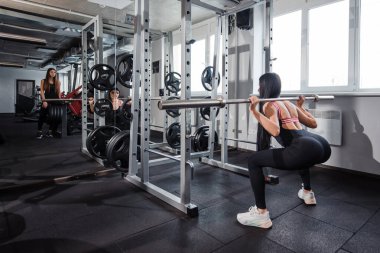 Aktif giyimli beyaz kadın modern spor salonunda halterle çömelme hareketi yapıyor. Siyah saçlı bir bayan arkadaş destek için yanında duruyor. Birlikte eğitim kavramı.